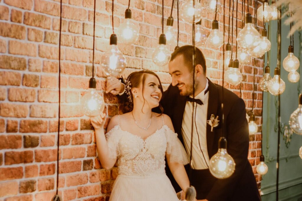 Photo of a Groom and a Bride Looking at Each Other Near Light Bulbs